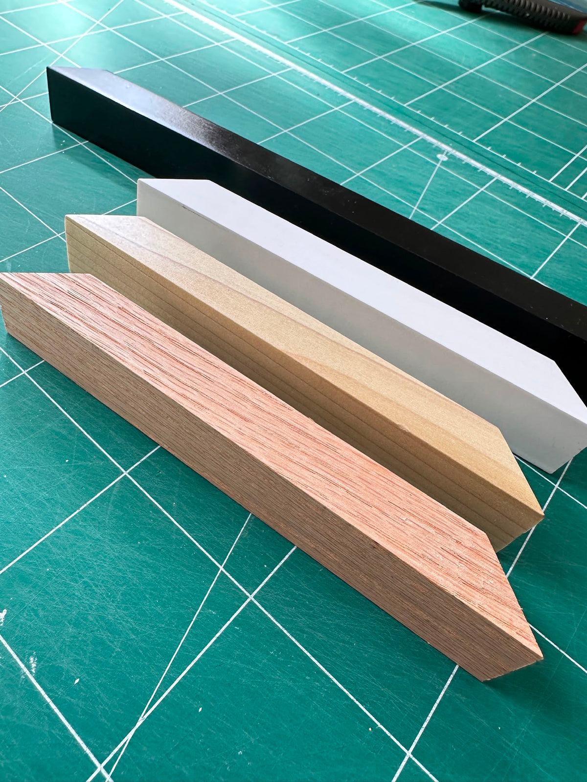 Four rectangular wood-look sample strips in different colors and finishes arranged diagonally on a green cutting mat with grid lines.