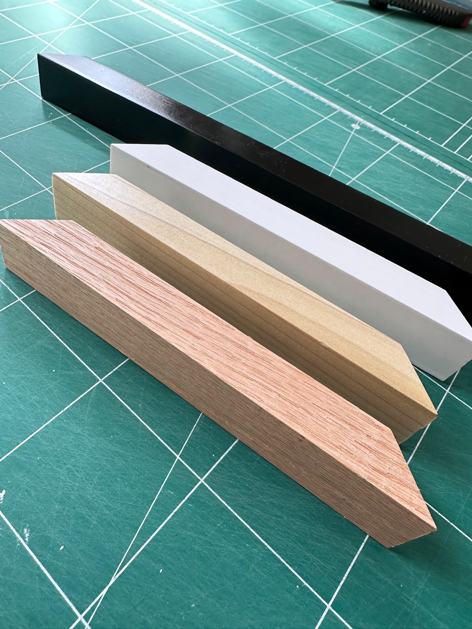 Four rectangular wood-look sample strips in different colors and finishes arranged diagonally on a green cutting mat with grid lines.