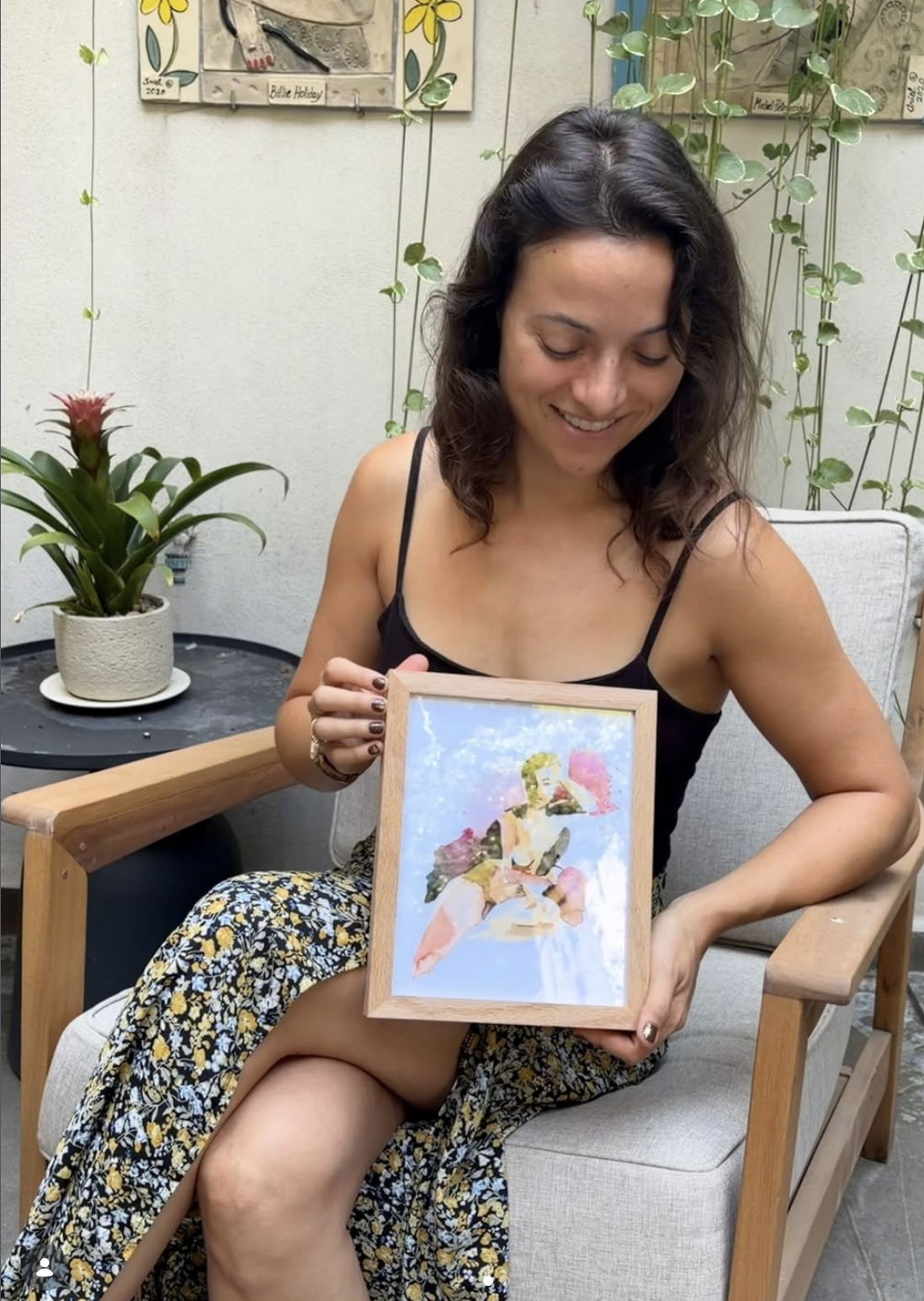 A woman sitting in a light gray armchair holds up a framed colorful painting of a bouquet of flowers. She is wearing a black spaghetti-strap top and a yellow and blue floral maxi skirt.