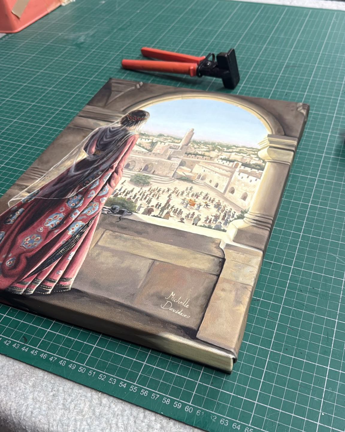 A rectangular canvas painting depicting a woman in a flowing red and dark robe looking out from an archway onto a city scene, placed on a green cutting mat on a workbench with tools nearby.