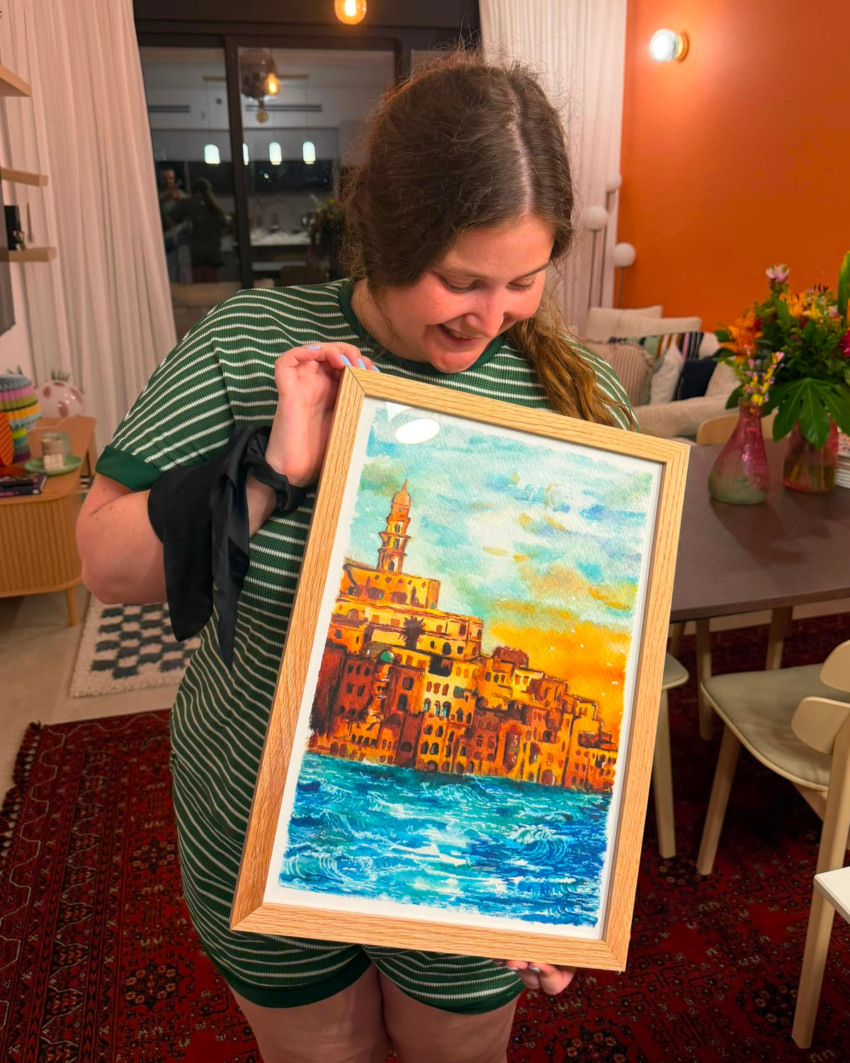A woman in a green striped shirt holds up a framed colorful painting of a coastal town with blue water and warm yellow-orange buildings, set in a home interior with an orange wall and dining table in the background.