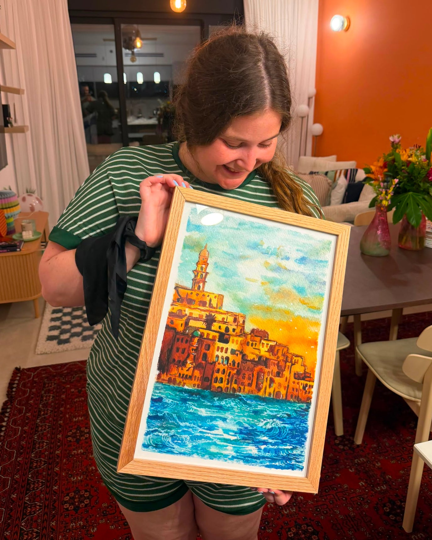 A woman in a green striped shirt holds up a framed colorful painting of a coastal town with blue water and warm yellow-orange buildings, set in a home interior with an orange wall and dining table in the background.