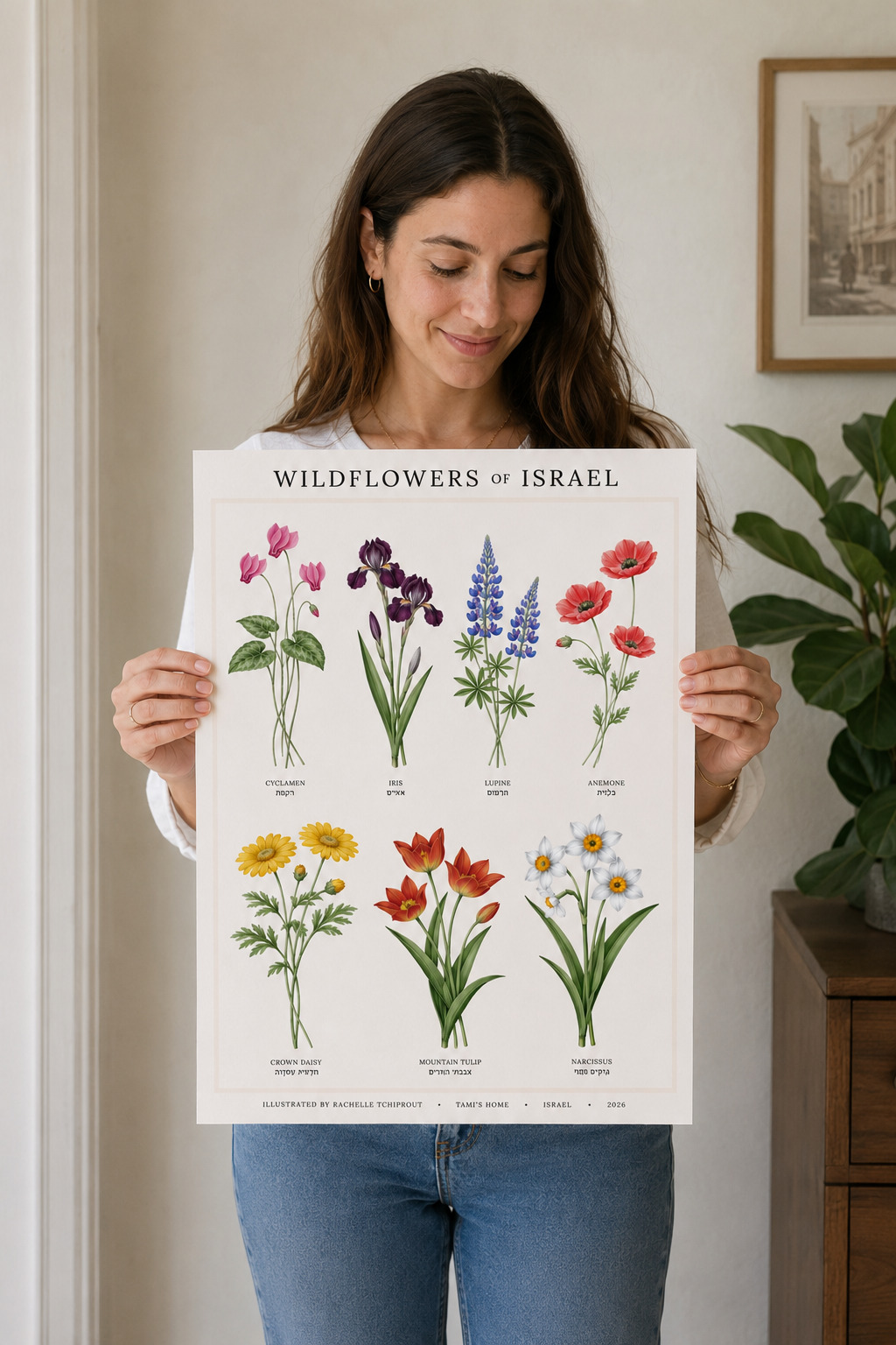 A woman holding a botanical art print titled 'Wildflowers of Israel' featuring various illustrated wildflower species. She is standing indoors in a home setting with a framed picture and a potted plant in the background.