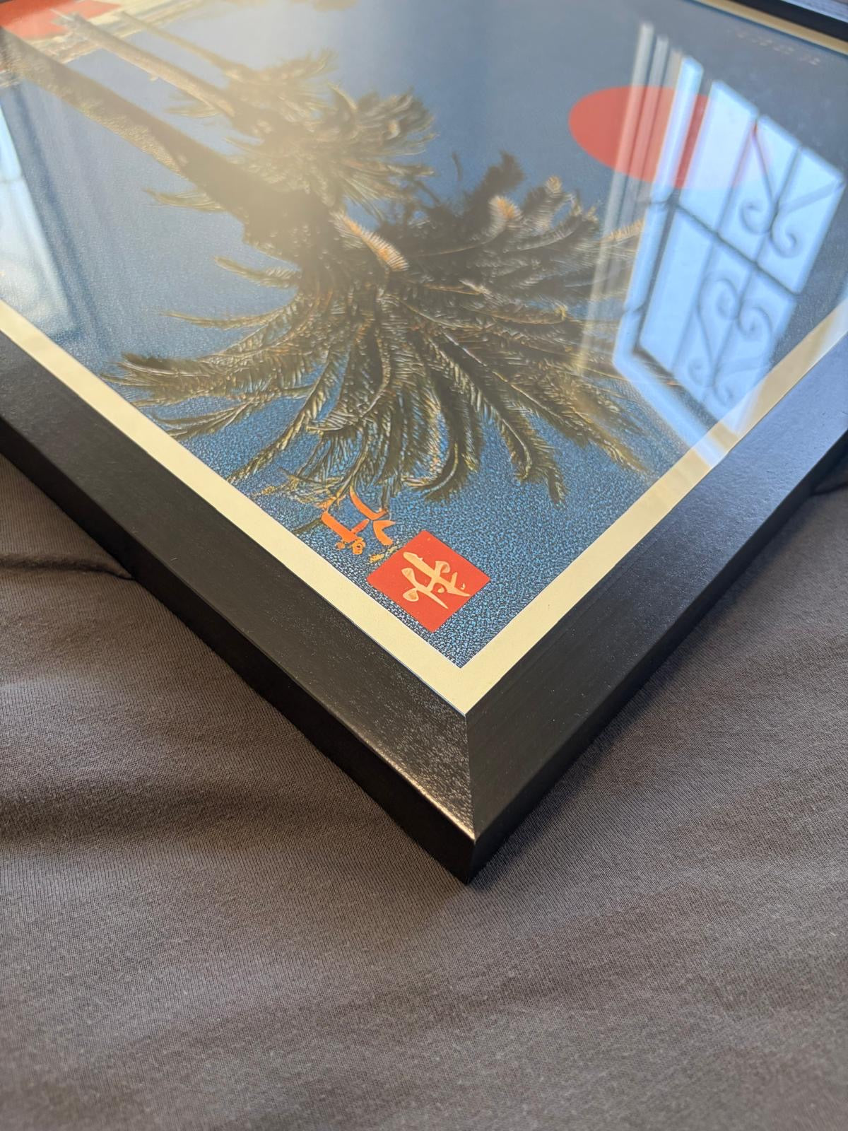 A framed art print featuring a Japanese-style design with a blue background, a black palm tree, a red sun disc, and red seal marks, shown at an angle on a gray fabric surface.
