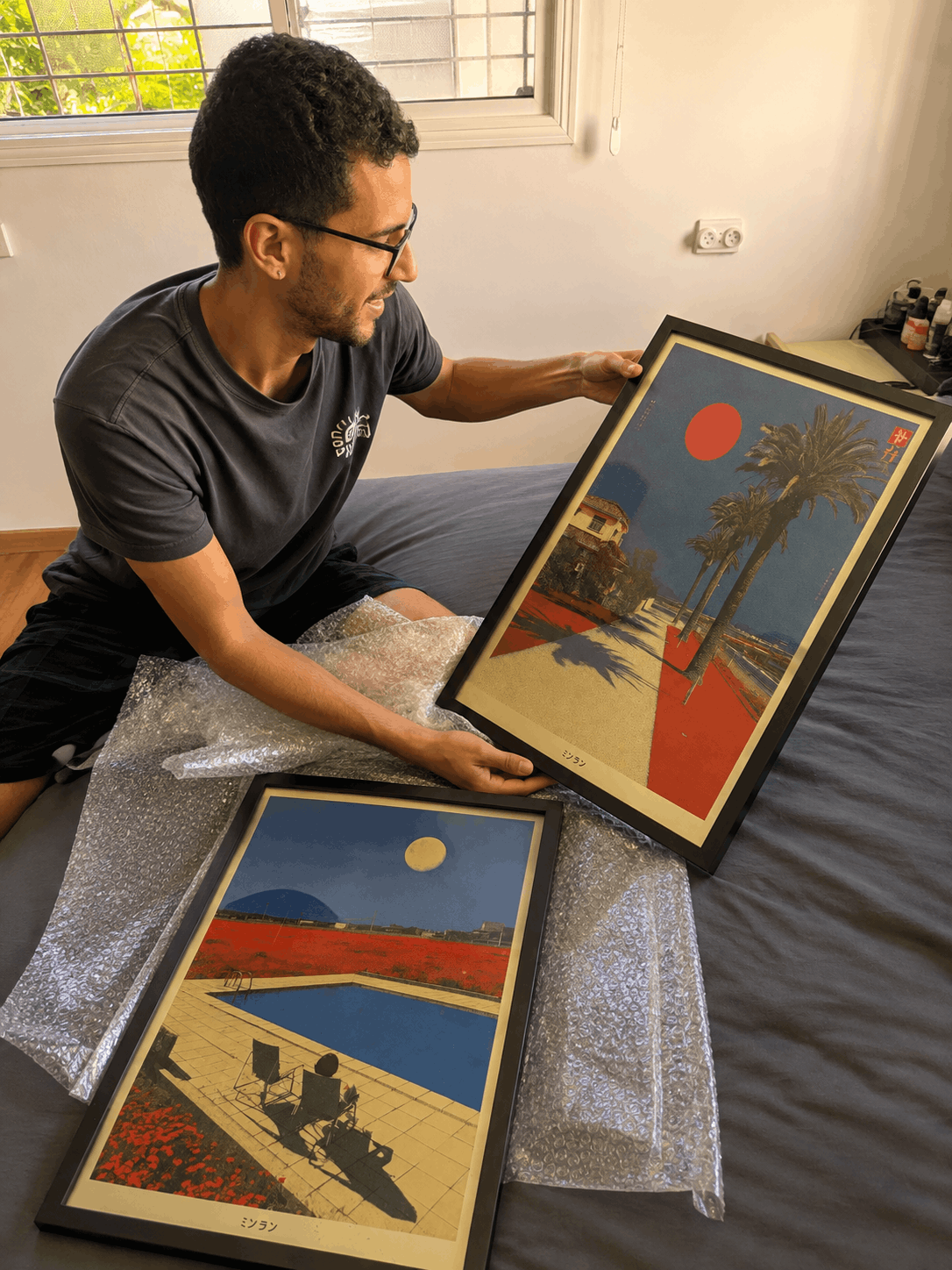 A man sitting on a bed unpacks two framed art prints using bubble wrap. The prints feature a bold, graphic, retro-inspired design with a red sun, blue sky, and palm trees.