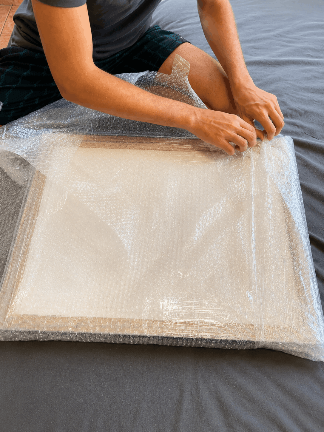 A person sitting on a bed is wrapping a large square framed item in clear bubble wrap. The frame appears light wood with a white or off-white mat, and the bed has dark gray bedding.